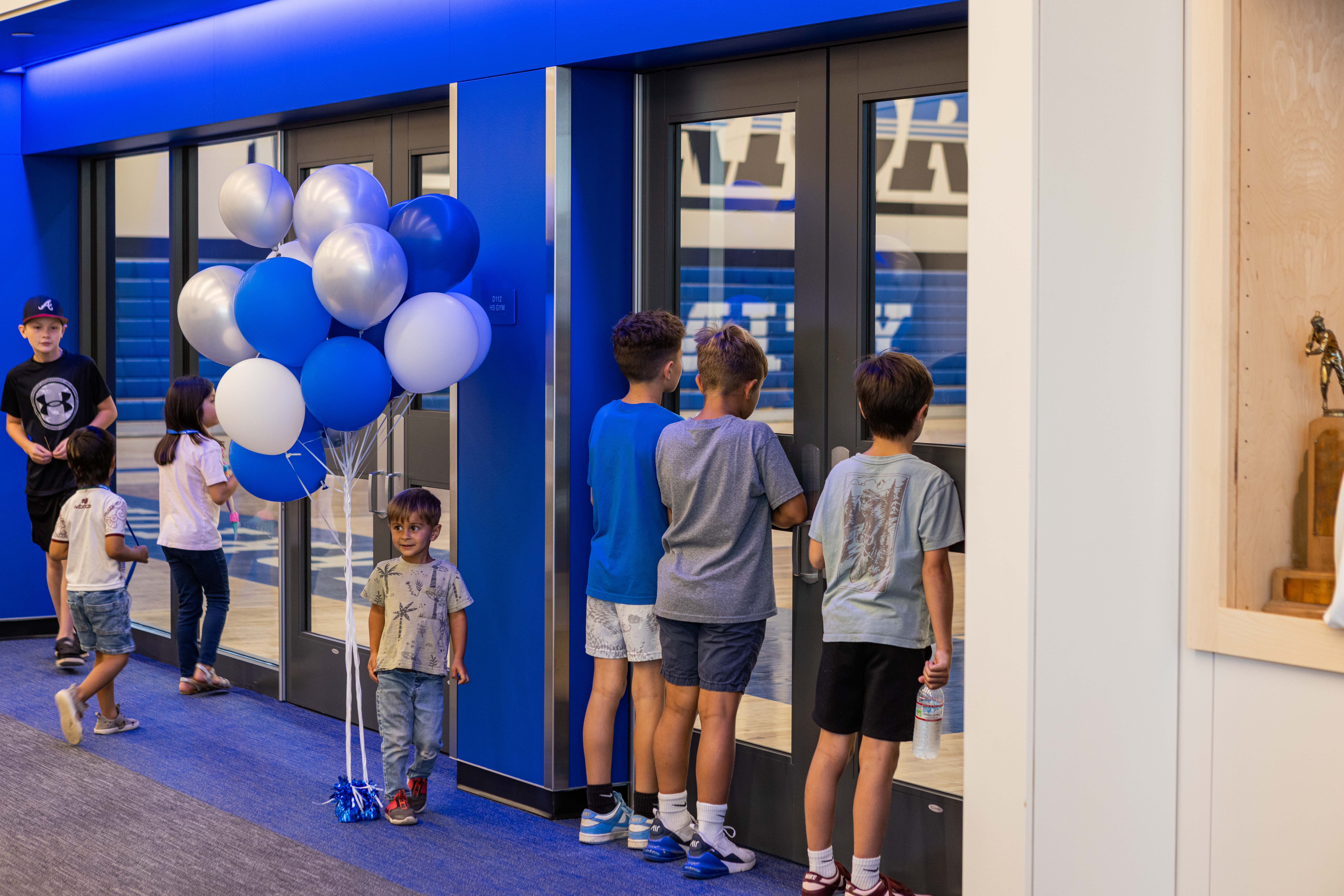 Inside the new entry corridor on opening day — six elementary-age kids waiting at the glass doors into the gym, a young boy holding a tall bunch of royal-blue and white balloons, royal-blue glowing wall behind them, a wood trophy case at right.