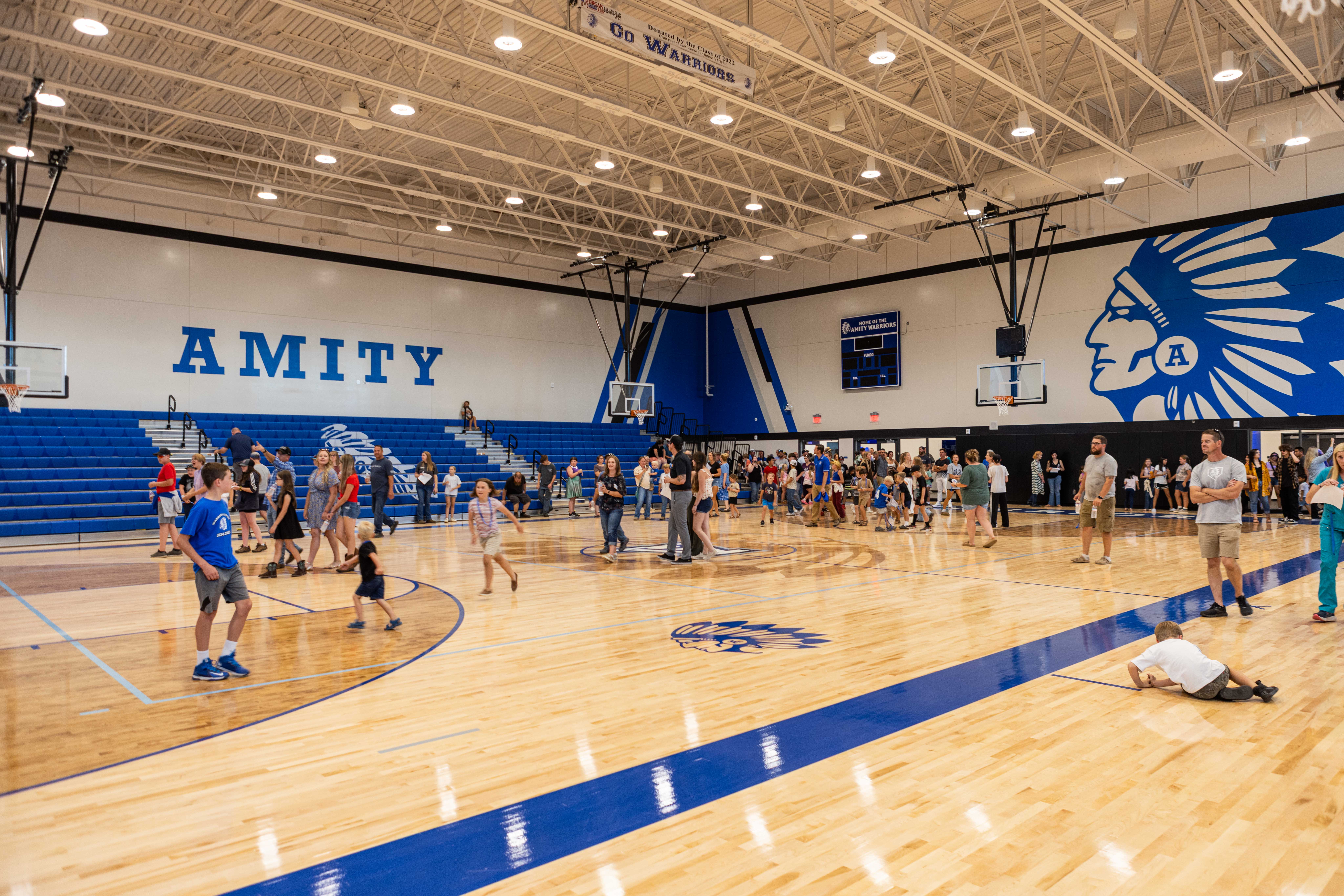 Inside the new Amity competition gym on opening day: a wide polished hardwood floor with the Warrior headdress logo and the AMITY wordmark large on the back wall, blue bleachers, families wandering the court, kids running, hanging banner reading 'Go Warriors'.