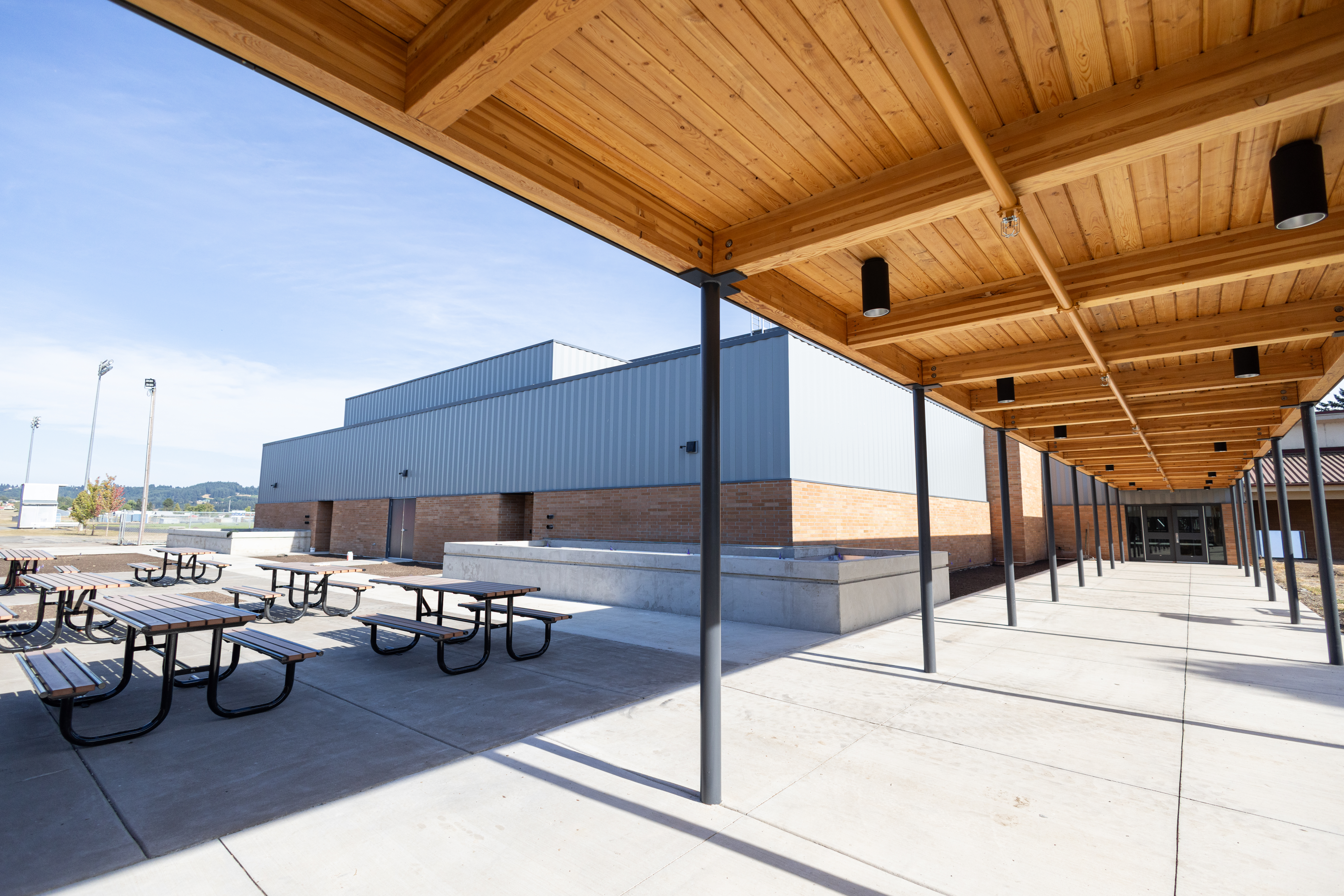 Outside the new building on a clear day — a long covered walkway with a warm timber-beam ceiling, brick columns supporting it, wood picnic benches under the cover, the new metal-and-brick athletic wing across the courtyard.