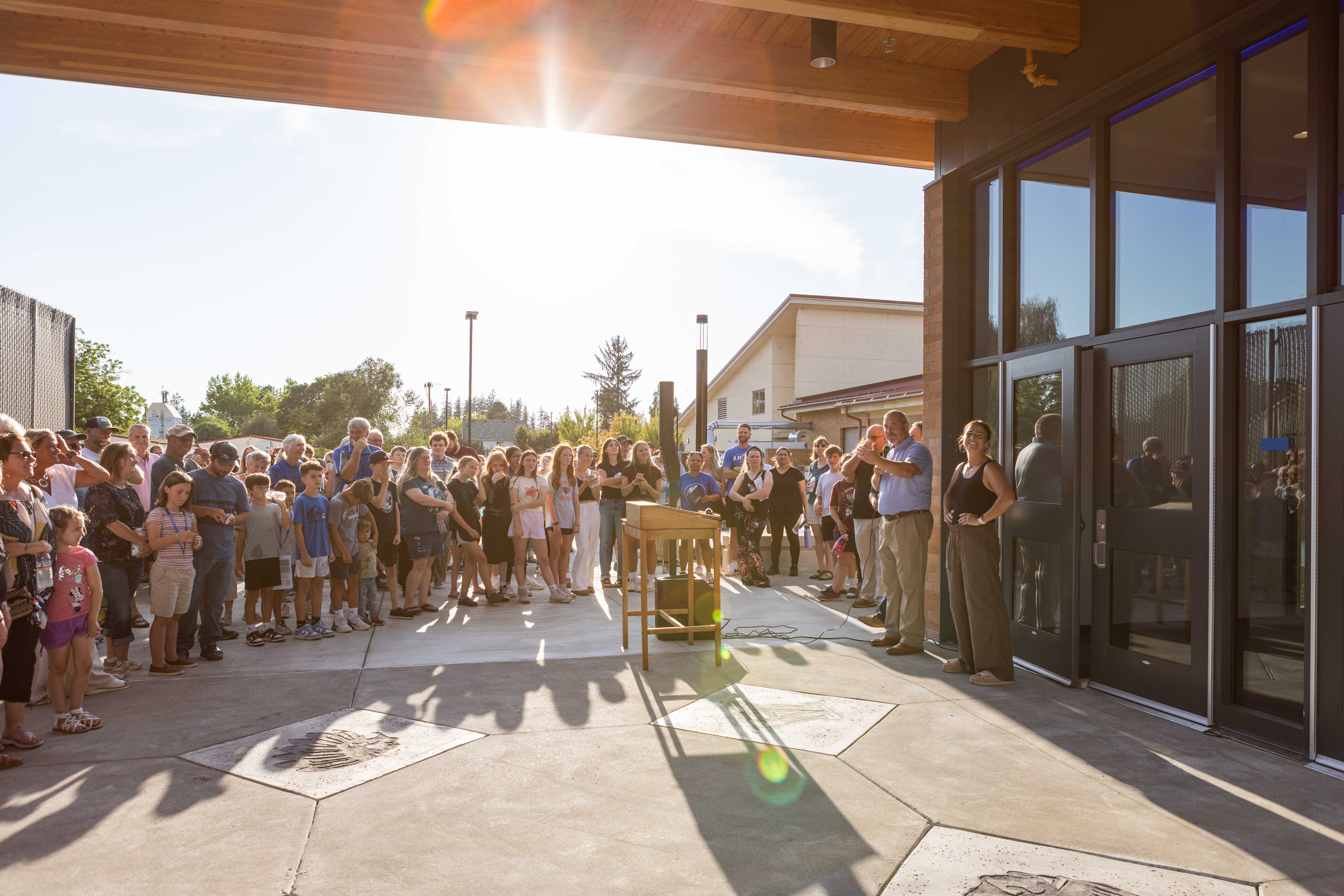 A crowd of Amity families and middle-school-age kids gathered for the outdoor opening ceremony at the new school entrance, late-afternoon sun streaming through the timber-beam roof overhead.