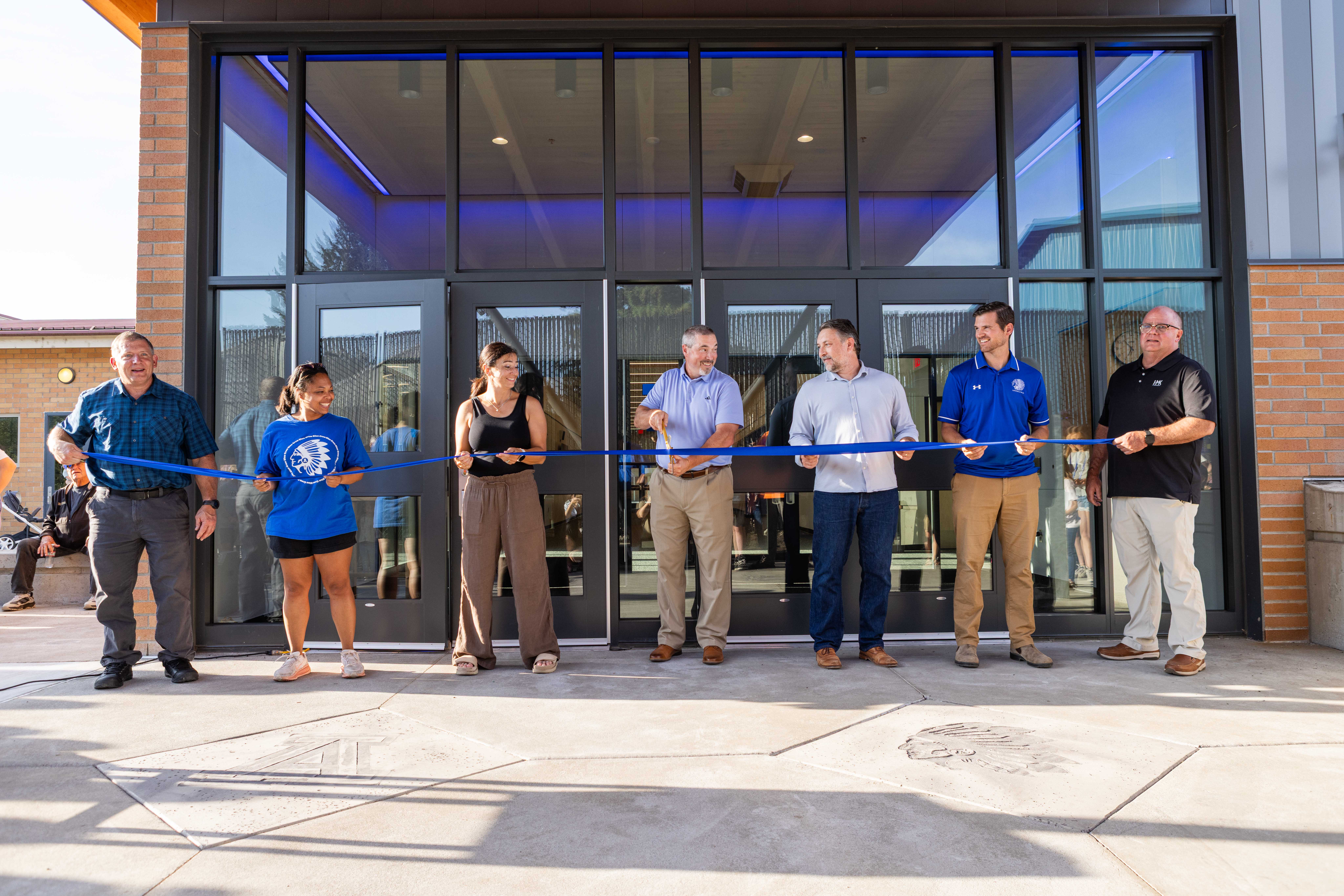 Seven community members stretching a blue ribbon across the front of the new glass entrance at Amity High School on opening day, the superintendent center-frame with scissors, a young Warrior staffer in the blue Warrior tee at left, the new lobby visible behind them.